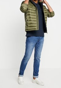 Olivově zelená prošívaná bunda se zipem vpředu, námořnická polo košile a modré džíny. Bílá teniska dokončuje outfit.