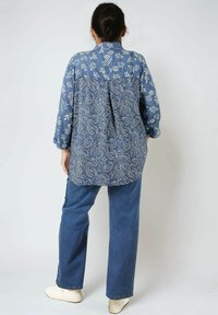Woman standing with back facing camera wearing blue floral and paisley patterned shirt, blue jeans, and white sneakers.