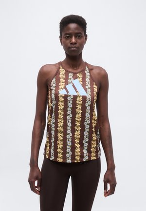 Kvinde iført brun ærmeløs top med lodrette gule og blå blomstrede striber samt stort lyseblåt Adidas-logo, stående mod ensfarvet baggrund.
