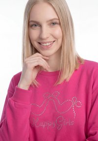 Blonde vrouw met steil haar die glimlacht, draagt een felroze sweatshirt versierd met kleine strass-steentjes en de woorden "Happy Girl".