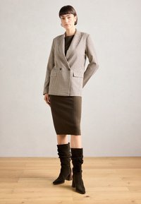 Sako s vzorem houndstooth, dvouřadové, olivová žebrovaná sukně a volné tmavozelené boty na podpatku s špičatými špičkami, umístěné na neutrálním pozadí.