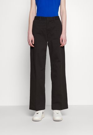 Person trägt gerade schwarze Hose, weiße Turnschuhe und ein blaues ärmelloses Oberteil, steht vor einem schlichten weißen Hintergrund.