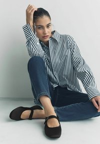 Une paire de chaussures Mary Jane noires avec une lanière et une boucle, associées à une chemise à rayures et un jean bleu, assises sur une surface claire.