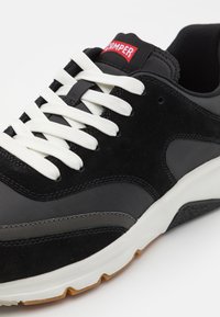 Schwarze Sneaker aus Leder und synthetischen Materialien. Mit weißen Schnürsenkeln, einer dicken Sohle und einem roten Logo-Tag auf der Zunge.