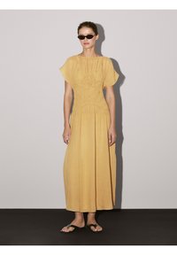 Maxi-jurk in mosterdgeel met geplooide textuur bij het lijfje, korte fladdermouwen en verticale plooien. Draag met zwarte sandalen.