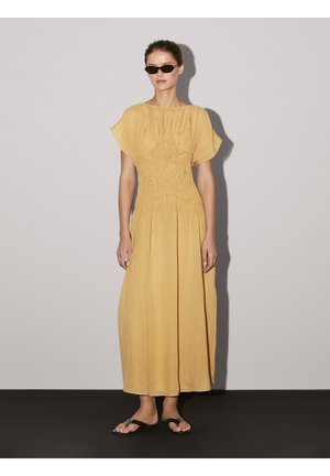 Robe maxi en jaune moutarde avec une texture froncée au niveau du corsage, des manches courtes en volants et des plis verticaux. Portée avec des sandales noires.