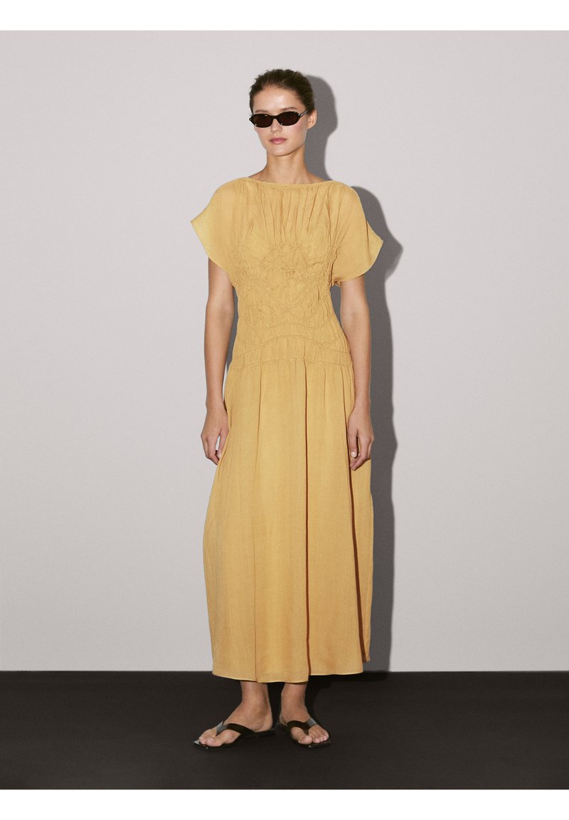 Maxi-jurk in mosterdgeel met geplooide textuur bij het lijfje, korte fladdermouwen en verticale plooien. Draag met zwarte sandalen.