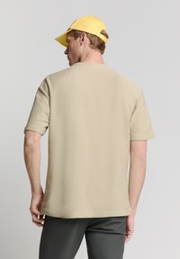 Beige t-shirt met korte mouwen en een gestructureerde afwerking, voorzien van een ronde halslijn. Model draagt een gele pet en staat met de rug naar de camera.