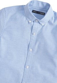 Camisa abotonada azul claro hecha de una tela con una textura sutil. Presenta un cuello tradicional y botones blancos a lo largo del frente.