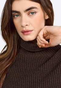 Brown ribbed turtleneck sweater with a soft texture. Model holds hand near face, showcasing natural makeup and long, wavy hair.