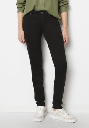 Person iført sorte slim-fit cargobukser med klaplommer, grøn langærmet skjorte og hvide sneakers, stående mod en hvid væg.