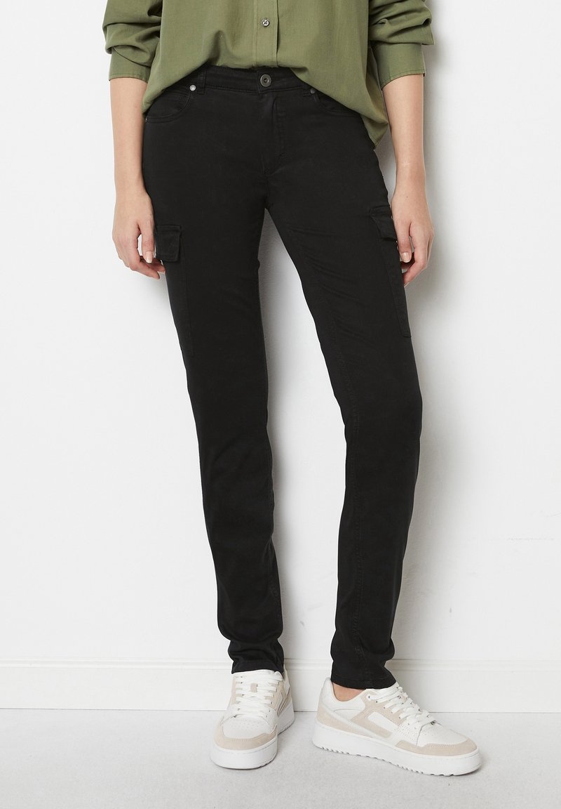 Personne portant un pantalon cargo noir slim avec poches à rabats, une chemise verte à manches longues et des baskets blanches, debout contre un mur blanc.