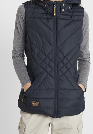 Persoon die een marineblauw doorgestikt mouwloos vest met voorzakken draagt over een grijs shirt met lange mouwen en beige cargobroek.