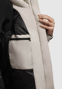 Light beige puffer jacket interior with black lining, featuring a zippered pocket and ribbed accents along the edges.