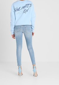 Sudadera azul clara con el texto "ACCIÓN ILIMITADA" en la parte posterior, combinada con jeans ajustados azul claros y sandalias de tacón a juego.