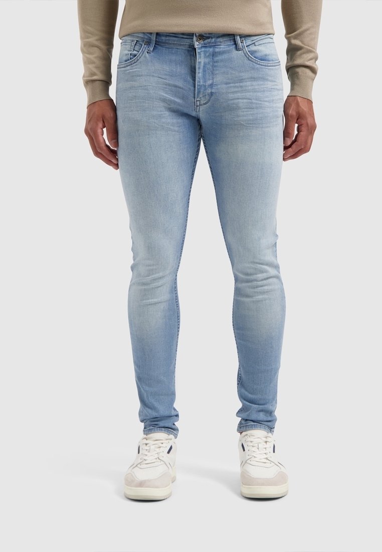 Man som bär ljusblå tajta jeans, beige tröja och vita sneakers, står mot en enkel ljusgrå bakgrund.