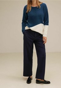 Kvinna i blå och vit färgblocktröja, marinblå vida byxor och svarta loafers, stående med händerna i fickorna.