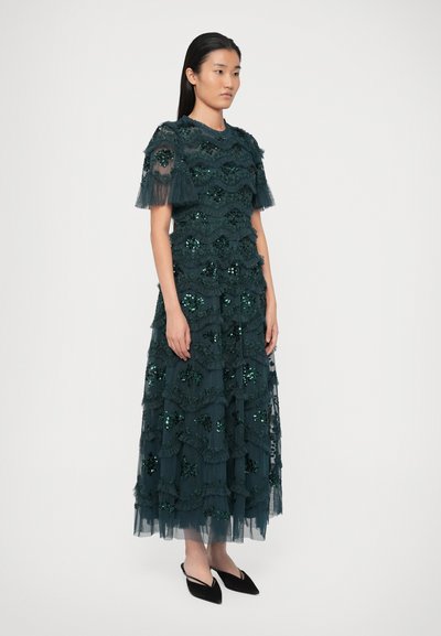 Teal, gestuftes Kleid aus transparenter Stoff mit Pailletten und Rüschen. Kurze Ärmel, runder Ausschnitt und knöchellanger Rock. Schwarze Ballerinas getragen.