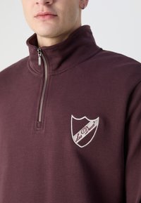 Bordo flis pulover s visokim ovratnikom i polu patentnim zatvaračem. Ima izvezeni logo u obliku štita u bijeloj i burgundi boji na prsima.