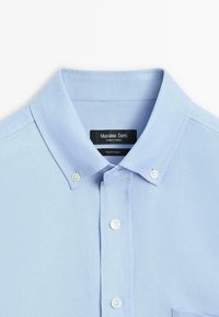 Light blue button-up shirt with a structured collar, made of textured fabric. Features white buttons and a chest pocket. Brand label visible.