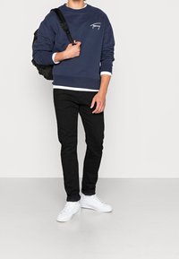 Man som bär en marinblå sweatshirt med vit logotyp, svarta skinny jeans, vita sneakers och bär en svart ryggsäck över ena axeln.