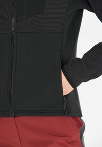 Veste noire avec panneau supérieur texturé, fermeture éclair à l'avant et poches latérales. Pantalons rouges contrastants visibles en dessous. Design simple et athlétique.