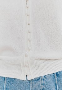 Cardigan blanc en tricot avec un ourlet côtelé, présentant de petits boutons texturés sur le devant. Associé à un denim bleu clair.