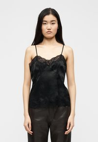 Νέα γυναίκα με μακριά σκούρα μαλλιά, που φοράει μαύρο σατέν καμισόλι με δαντέλα και σκούρα δερμάτινα παντελόνια, στεκόμενη μπροστά σε απλό φόντο.