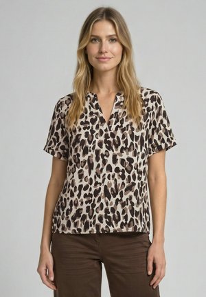 Vrouw met lang blond haar die een beige blouse met korte mouwen draagt met zwarte en bruine luipaardvlekken en een bruine broek, staand tegen een lichte achtergrond.
