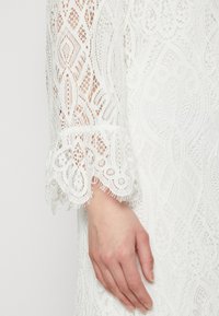 Bela čipkasta obleka z zapletenimi vzorci, razširjenimi čipkastimi manšetami in teksturiranim zaključkom, ki prikazuje nežne podrobnosti in eleganten dizajn.