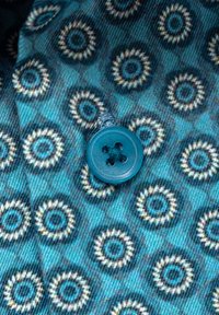 Blauwe stof met cirkelvormige patronen in wit en blauw rondom een centraal geplaatste blauwe knoop met vier gaten.