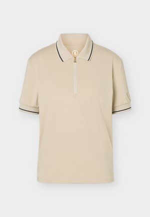 Joven de pie mirando hacia adelante, vistiendo un polo beige de manga corta, pantalones negros con cinturón y zapatillas blancas sobre un fondo claro.