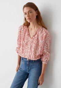 Blusa leggera e modellata con motivo floreale rosso su uno sfondo chiaro, caratterizzata da un collo a V e maniche arricciate, abbinata a jeans blu.