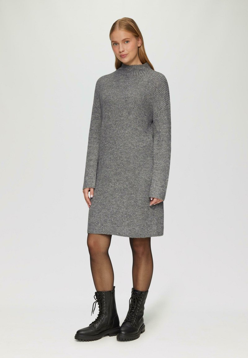 Graues Strickkleid mit hohem Kragen, langen Ärmeln und gerader Passform, kombiniert mit schwarzen Schnürstiefeln im Kampfstil. Glatte Textur über das gesamte Kleidungsstück.