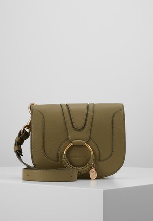 Olijfgroene leren crossbodytas met gouden ringdetail en verstelbare band, geplaatst op een wit oppervlak tegen een grijze achtergrond.