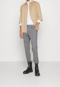 Uomo che indossa una camicia beige a maniche corte con bottoni sopra un dolcevita bianco, pantaloni grigi a scacchi e stivaletti neri in pelle, in piedi su un pavimento grigio.