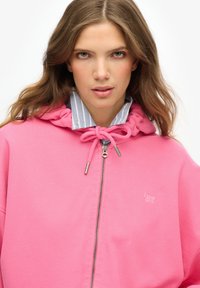 Haut à zip rose en tissu doux, doté d'une capuche avec cordon de serrage et d'une fermeture éclair argentée. Un t-shirt rayé bleu clair est visible en dessous.