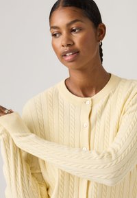 Νεαρή γυναίκα με σκούρα μαλλιά και σκουλαρίκια, φοράει ένα παλ παστέλ κίτρινο πλεκτό ζακέτα με κουμπιά, κοιτάζοντας προς τα πλάγια με το χέρι σηκωμένο.