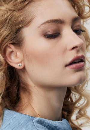 Złote kolczyki-stud z okrągłymi przezroczystymi kamieniami szlachetnymi, osadzone w delikatnym wzorze. Modelka ma falujące blond włosy i nosi jasnoniebieską bluzkę.