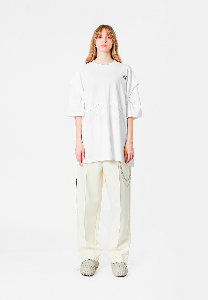 Camiseta blanca de gran tamaño con mangas cortas y cortes únicos. Combinada con pantalones anchos crema con detalles en cadena y calzado gris texturizado.