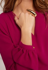 Fioletowa bluzka z dekoltem w serek i bufkami. Złoty, metalowy pierścień na dłoni, prezentujący gładką teksturę i zaokrąglony kształt.