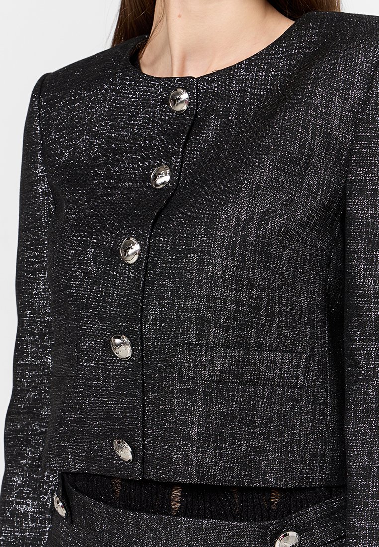 Blazer noir texturé avec des boutons en argent, longueur courte, et une petite poche sur le devant. Des fils métalliques créent un léger scintillement.