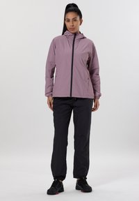 Lätt, lavendelfärgad vattentät jacka med huva, frontdragkedja och sidofickor, i kombination med svarta byxor och svarta träningsskor.