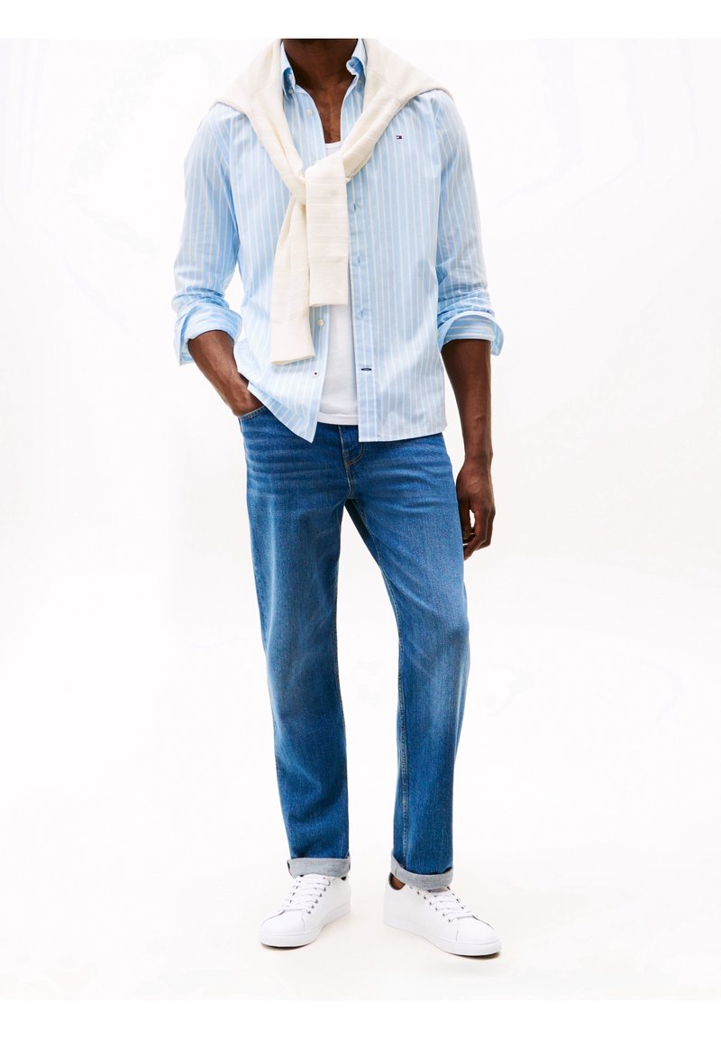 Personne portant une chemise bleue à rayures avec un pull blanc drapé sur les épaules, un jean bleu retroussé aux chevilles et des baskets blanches, debout avec une main dans la poche.