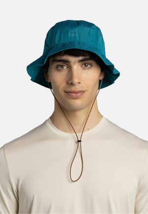 Joven con un sombrero tipo pescador de color verde azulado con correa ajustable para la barbilla y una camisa beige clara, mirando hacia adelante con expresión neutral.