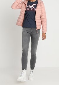 Person klädd i en rosa puffjacka, en marinblå Hollister-t-shirt, grå skinny jeans och vita snörskor, står mot en neutral bakgrund.