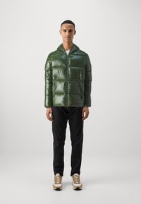 Veste rembourrée verte avec une finition brillante, dotée d'une fermeture éclair, d'une capuche et de coutures carrées. Associée à un jean sombre et des baskets de couleur claire.