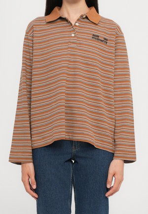 Personne portant un polo à manches longues rayé marron et blanc avec col, et un jean bleu, debout devant un fond blanc uni.