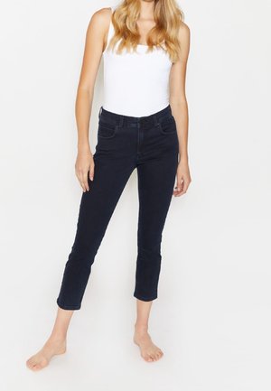 Vrouw staat blootsvoets, draagt een wit mouwloos topje en zwarte cropped skinny jeans tegen een effen witte achtergrond.