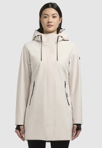 Manteau imperméable beige clair avec capuche, doté de poches zippées noires, d'un design épuré et de manches longues avec des poignets intérieurs pour plus de chaleur.